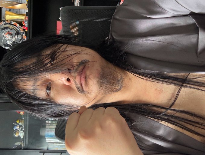 A close-up selfie of a young man with long dark hair and a light beard, wearing a dark top, photographed in what appears to be an arcade setting with game cabinets visible in the background.