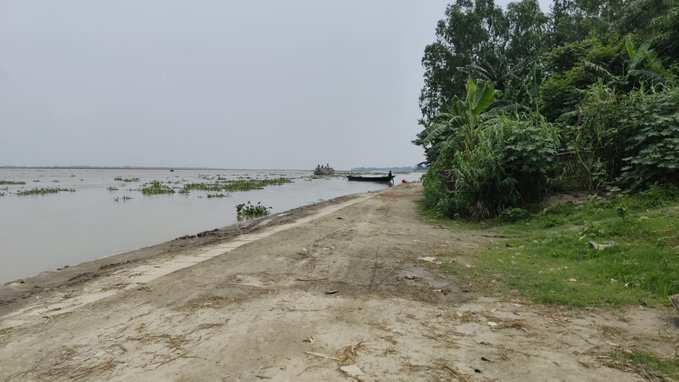 A dirt road runs along a riverbank, with boats moored in the distance and lush green vegetation and banana trees lining the right side under an overcast sky.
