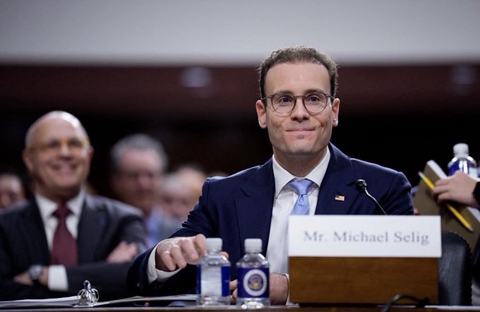 A man in glasses and a navy suit smiling at a congressional hearing, with a nameplate reading 'Mr. Michael Selig'.