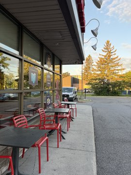 An empty outdoor patio area with black tables and red chairs sits in front of a building with large windows, alongside a parking lot with a van and trees under a partly cloudy sky.