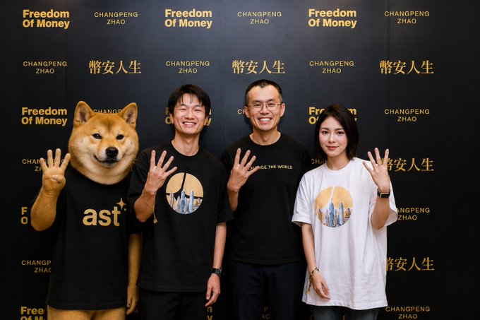 Three people and a person in a Shiba Inu costume pose in front of a backdrop displaying "Freedom Of Money" and "CHANGPENG ZHAO", with the Shiba wearing an "ast" t-shirt.