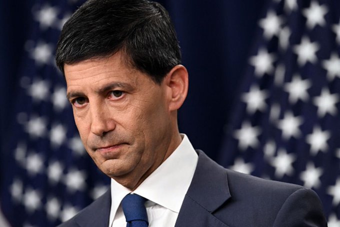 A serious-looking man in a dark suit and blue tie stands before a backdrop of American flags. The image likely depicts former Federal Reserve Chair nominee Kevin Warsh, referenced in the post as the Fed Chair whose Jackson Hole speech could drive major market moves.