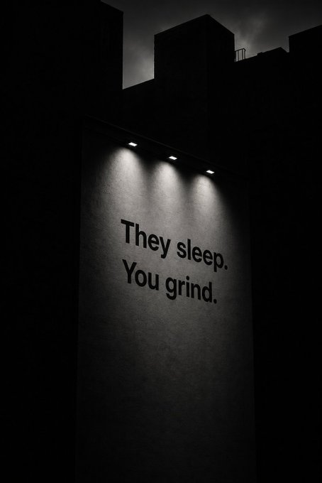 A dramatic nighttime billboard reads "They sleep. You grind." in bold white letters, illuminated by three overhead spotlights against a dark sky and silhouetted buildings. The image reinforces the post's message about relentless dedication, portraying the grind as a defining trait that separates committed traders from the rest.