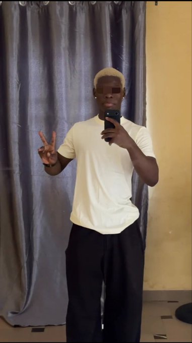 A mirror selfie of a young man with bleached blonde hair wearing a white t-shirt and black pants, making a peace sign in front of gray curtains.