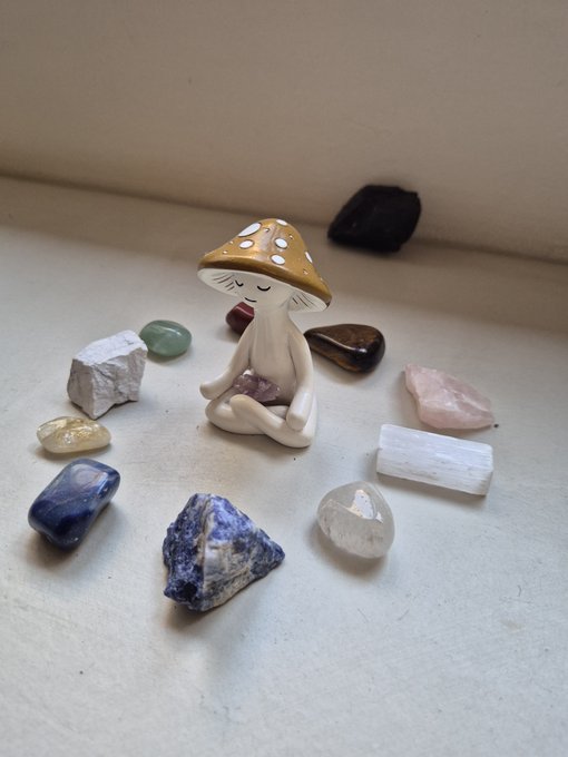 A small ceramic meditation figurine of a girl with a mushroom cap for a head, holding an amethyst, surrounded by an assortment of colorful healing crystals on a white surface.