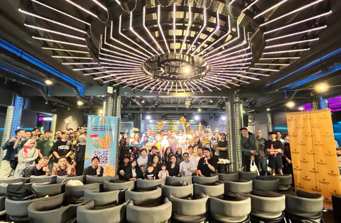 The image shows a large group of people gathered together at an event or conference, possibly a music festival or a tech convention. The attendees are standing around each other, posing for photos, and some are holding up signs. There are several chairs placed throughout the scene, indicating that this is likely a venue with seating arrangements for guests.

The post appears to be related to the event, as it mentions terms like "Makasih Bandung frens DeFi sessions," "Web3 alpha," "merch," and "packed house." These words suggest that the image might have been taken at an event where these topics are being discussed or showcased. The attendees seem to be enjoying themselves and engaging in conversations, which further supports the idea of a social gathering or conference.