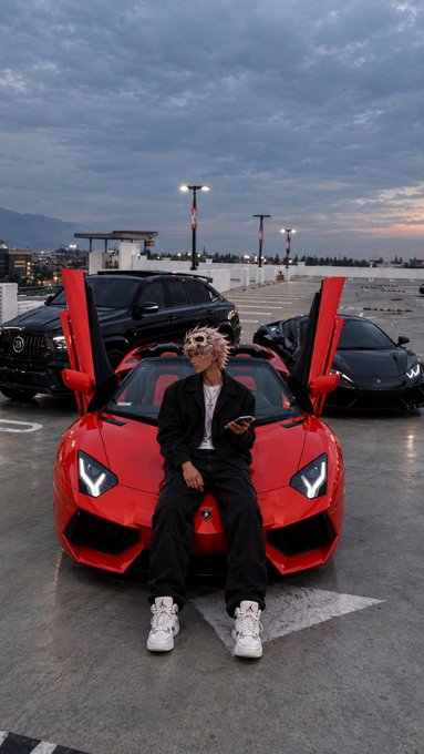 A young person with pink hair sits on the hood of a bright red Lamborghini with its scissor doors open, holding a phone in a parking lot at dusk with other luxury vehicles behind.