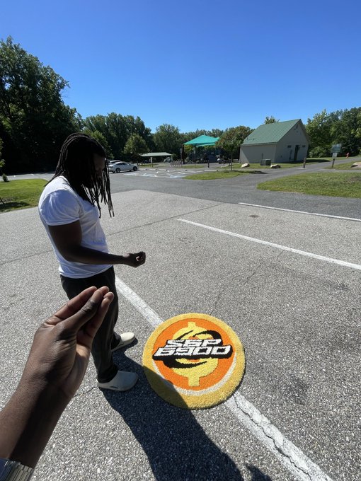 The image features a person with dreadlocks standing next to a yellow and orange circular sign that reads "Day 241 of posting this rug until SPX690 0 flips the stock market". The person appears to be walking towards or away from the sign, possibly indicating their thoughts on the current state of the stock market.

There are several cars parked in the background, suggesting an urban setting. Additionally, there is a parking lot with multiple cars and a truck visible in the scene.