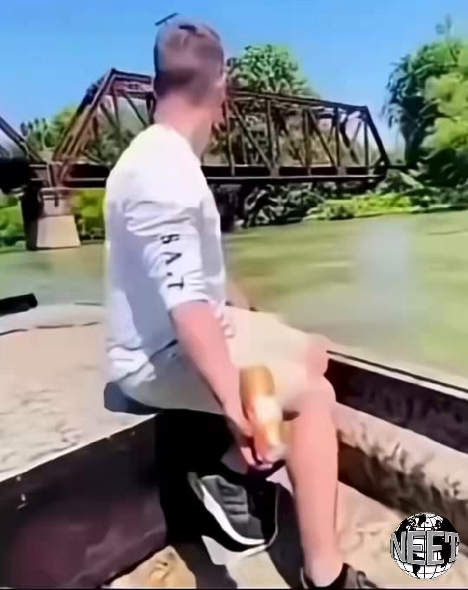 The image shows a man sitting on a bench near a river, holding a beer bottle. The scene appears to be set during the day with a bridge visible in the background. There are also some text elements present in the image that seem unrelated to the main subject of the photo.