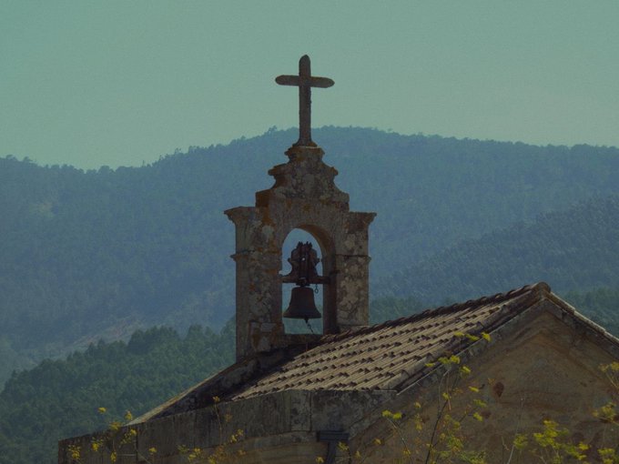 A weathered stone bell tower topped with a cross stands against hazy mountains at dusk, rising above a tiled roof.