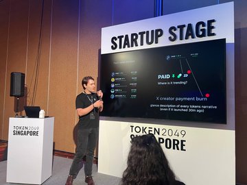 A speaker presents on the "STARTUP STAGE" at a "TOKEN2049 SINGAPORE" event, standing next to a large screen showing a payment burn chart.