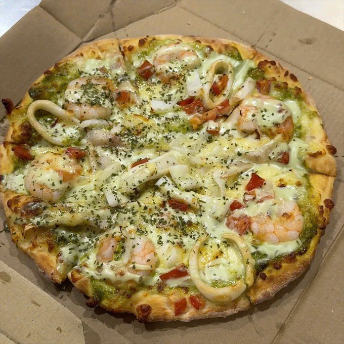 A freshly baked seafood pizza with shrimp, calamari rings, and melted cheese on a green pesto base, sitting inside a cardboard delivery box.