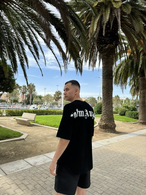 A young man in a black graphic t-shirt and dark shorts walking along a paved path lined with tall palm trees on a sunny day.