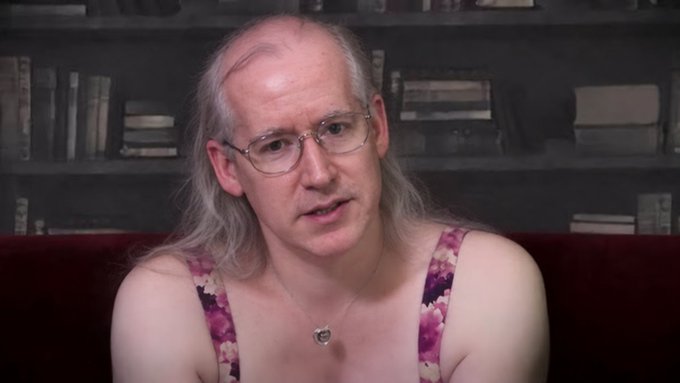 A person with long grey hair and glasses wears a floral pink and purple camisole top while sitting on a red couch in front of bookshelves.