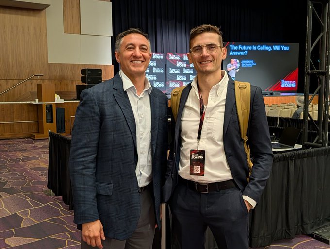 Two men in suits pose together at a Money 2020 conference, with a screen behind them reading 'The Future is Calling, Will You Answer?'