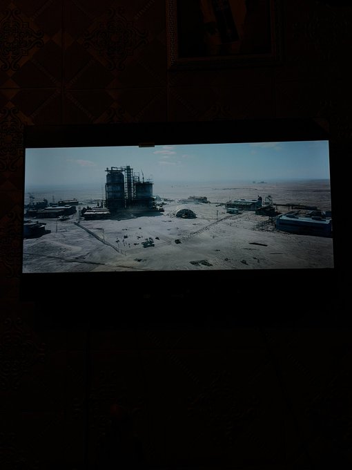 A screen in a darkened room displaying a video game or cinematic scene of an industrial facility or power plant in a desert landscape with vehicles parked outside.