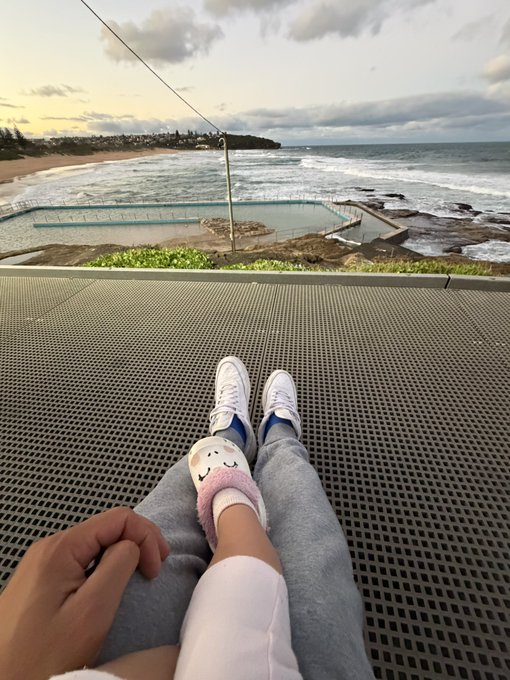 A first-person view of someone wearing white sneakers, gray sweatpants, and cute fuzzy slippers, relaxing on a metal grate overlooking an ocean tidal pool and beach at sunset.