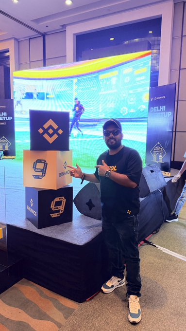 A man in sunglasses and a black t-shirt poses in front of a Binance booth at a 'Delphi Meetup' event.