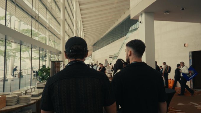 Two men in black shirts and a backwards cap walk through a spacious, sunlit convention hall or expo center, with other attendees visible in the background and a partially legible "AM" sign on a display to the right—evoking the energetic atmosphere of a GM-branded industry event or trade show.