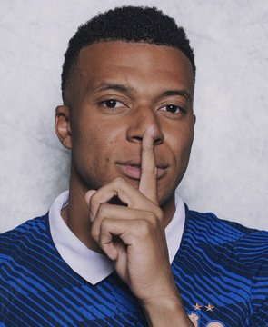 A close-up portrait of soccer player Kylian Mbappé in a dark blue striped jersey, holding his index finger to his lips in a 'shush' gesture.