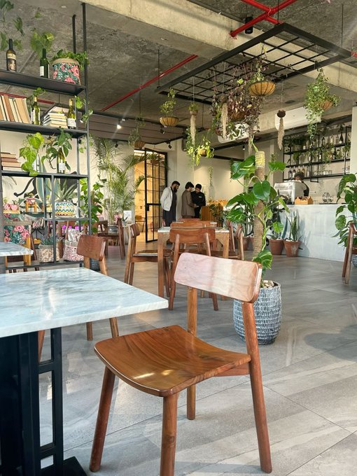 A bright, plant-filled café interior featuring wooden chairs, marble-topped tables, bookshelves, hanging potted plants, and a few people standing near the counter in the background.