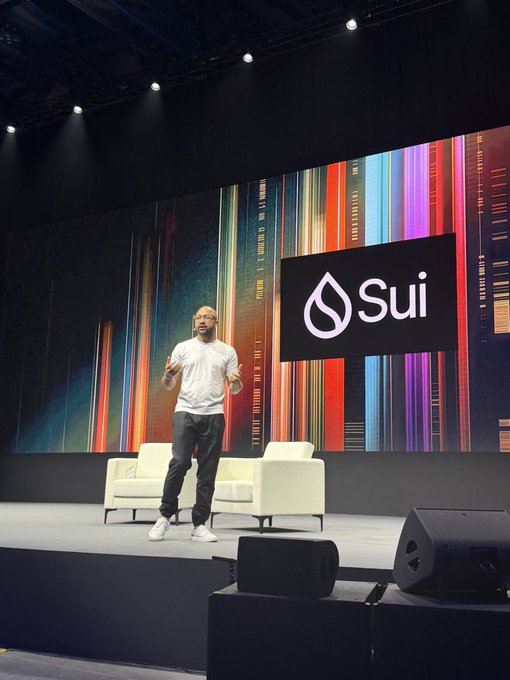 A man wearing a white t-shirt stands on a stage giving a presentation, with a large screen behind him displaying the 'Sui' logo and colorful vertical stripes.