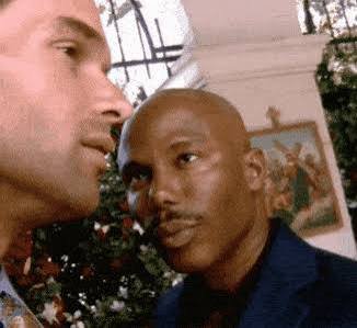 A close-up still shows two men outdoors, one in profile at the left and a bald man at right looking toward him, with a building and greenery behind them.