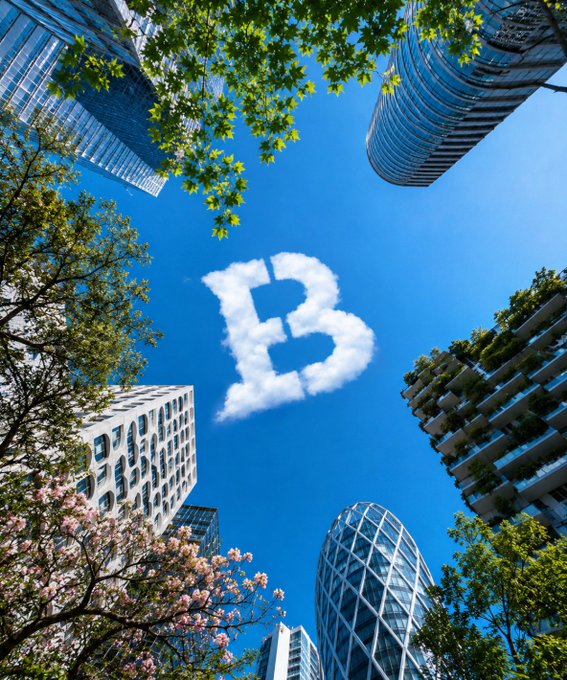 A cloud shaped like the letter "B" floats in the blue sky above modern skyscrapers and blooming trees, symbolizing the Biconomy brand featured in the "GM" crypto post. The cloud-formed "B" serves as a creative visual pun for Biconomy, tying the cheerful morning greeting to the project's identity.