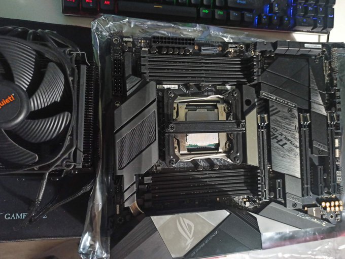 A computer motherboard, still partially in its anti-static packaging, sitting next to a be quiet! CPU cooler fan.