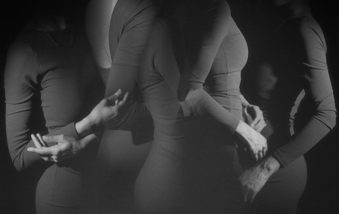 A dramatic black and white photograph captures three figures wearing tight-fitting, long-sleeved garments, with overlapping ghostly silhouettes created by motion blur. The monochromatic palette emphasizes the interplay of light and shadow on the contoured forms, reinforcing the artistic, noir-inspired aesthetic suggested by the "QT your black & white" caption.