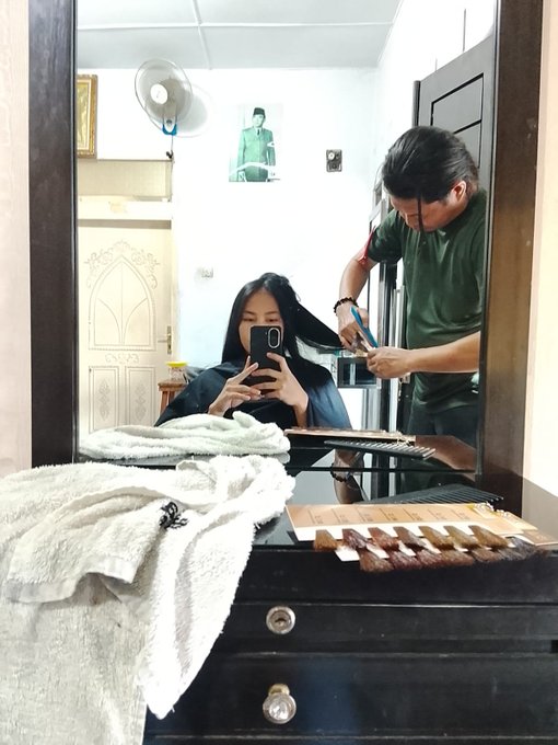 A mirror selfie captures a woman getting her long dark hair cut by a hairstylist in a green shirt, with hair samples displayed on a dresser and a fan on the wall behind them.