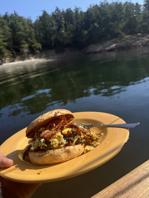 A breakfast sandwich with bacon, eggs, and cheese sits on a yellow plate held up against a scenic backdrop of calm water and a forested shoreline.