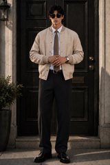 A stylish man standing in front of a dark doorway, wearing sunglasses, a cream-colored varsity jacket over a white shirt and brown tie, paired with black trousers.