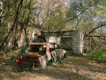 An abandoned, rusted vintage car overgrown with vegetation in front of a dilapidated concrete building in a wooded area.