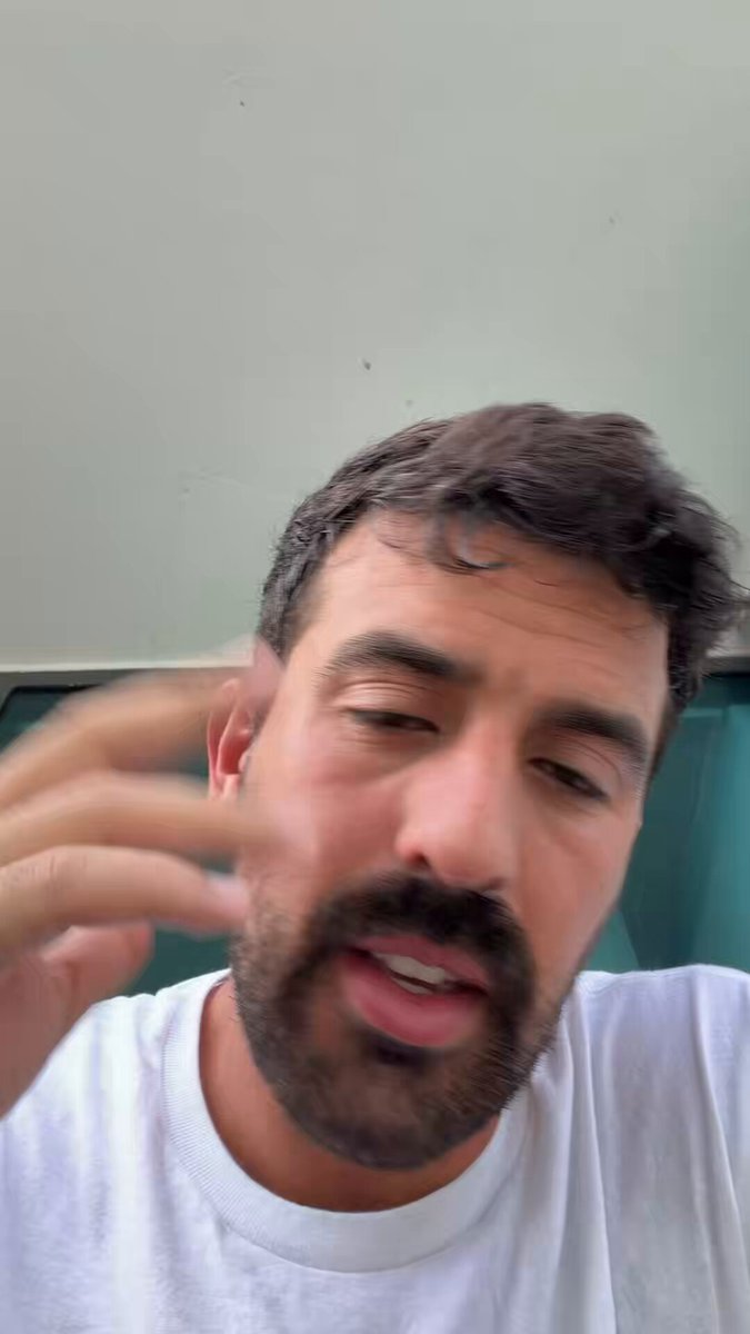 A man with a facial beard wearing white t-shirt is standing close to a wall, scratching his head. His expression suggests that he may be puzzled or concerned about something related to an article of clothing (a shirt) displayed on the right side of the image in the form of a caption.