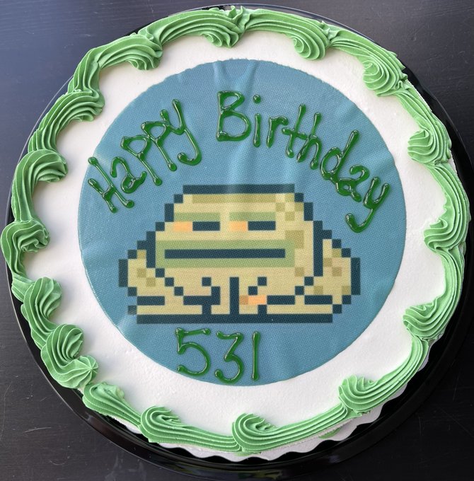 A round birthday cake features a pixelated green frog (Cryptoadz NFT character) printed on top, surrounded by a decorative green frosting border, with "Happy Birthday" written in green icing and "531" below the frog. The cake celebrates the 5th birthday of the Cryptoadz NFT collection, with the "531" likely referencing the collection's token ID or contract.