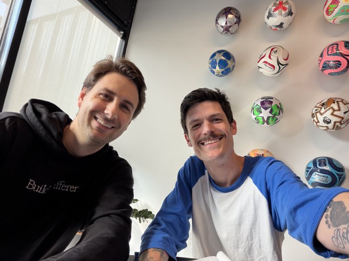 A selfie of two smiling young men, one wearing a black hoodie that says 'Build Differer', sitting indoors with a wall decorated with various colorful soccer balls behind them.