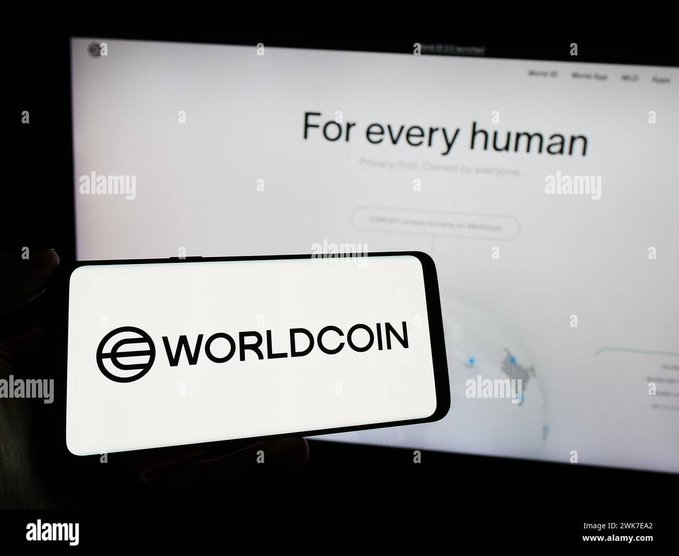 A smartphone displaying the "WORLDCOIN" logo is held up in front of a monitor showing the World website with the headline "For every human," visually representing the Worldcoin ecosystem discussed in the post about World Chain's scaling developments.