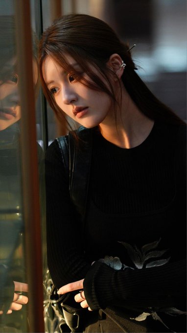 A young woman with dark hair wearing a black top poses for a portrait, with sunlight illuminating her face and a faint reflection visible on the left.