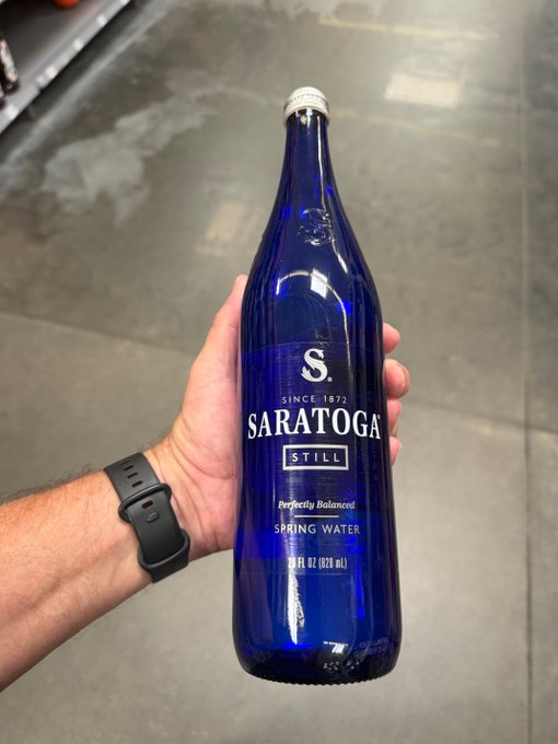 A hand holds up a 28 fl oz (828 mL) cobalt blue glass bottle of Saratoga Spring Water (Still, "Perfectly Balanced"), suggesting it's a premium bottled water often seen at conferences and events despite its upscale branding.