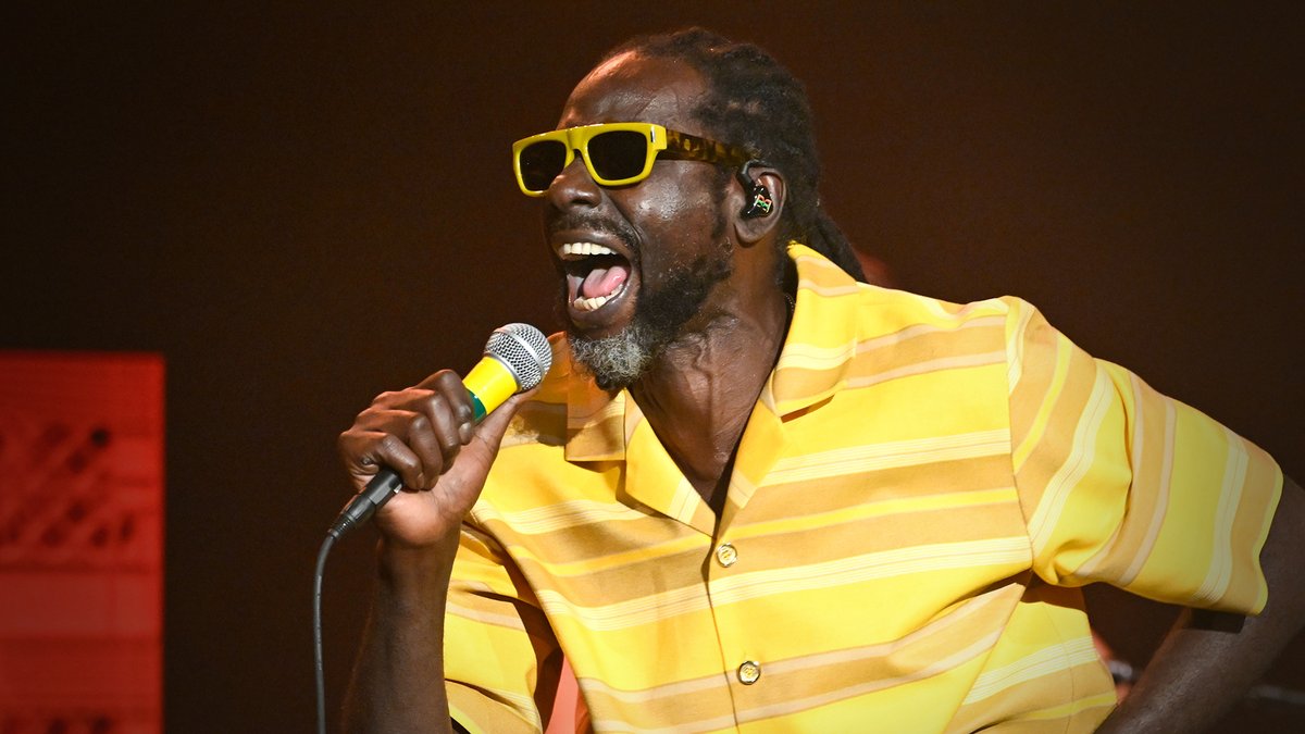 A concert photo of a Black male singer with dreadlocks wearing yellow sunglasses and a yellow striped shirt, singing passionately into a microphone.