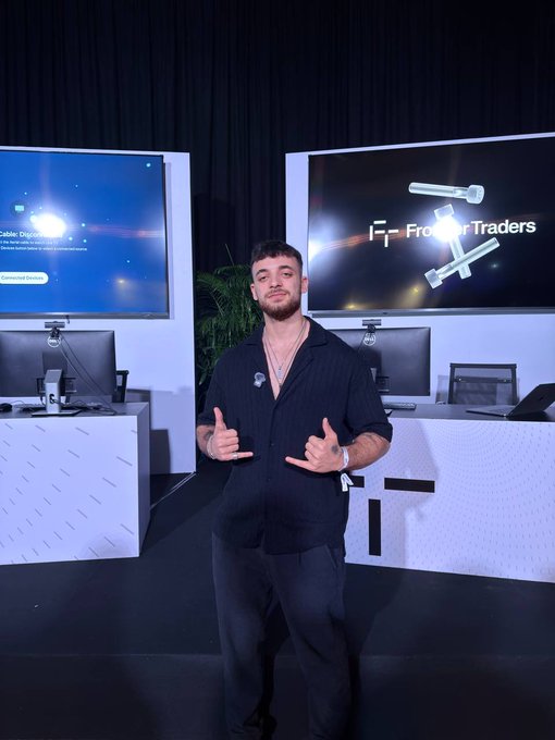 A young man poses in front of a "FrontRunner Traders" display, making hand gestures as part of a "GM" (good morning) greeting. The setup features screens showing trading platform login prompts and a gavel graphic, suggesting a trading-related event or meetup.