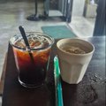 Two iced drinks in plastic cups, one dark (likely iced coffee or cola) and one light brown (likely iced latte), with straws on a dark wooden table.