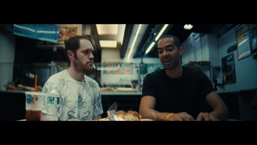 The image shows two men sitting at a table with food in front of them. One man has a water bottle next to him while they eat their meal together. The scene takes place in an indoor setting, possibly a restaurant or cafe, as indicated by the presence of a menu on one side and a TV screen visible in the background. A caption above the image reads "As cash declines, so does our ability to transact outside the evil eye". This suggests that the image might be related to a movie or TV show featuring characters who are affected by the decline of cash circulation in society.