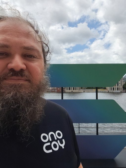 The image features a man with a beard wearing a black shirt, standing next to a large sculpture or art piece that has a blue and green color scheme. The man appears to be posing for a photo while looking directly at the camera. The background of the image shows a body of water, possibly an ocean, which adds to the overall atmosphere of the scene. The post seems to be related to the Solana Summit held in Berlin, as it mentions that the image was taken during the previous day's summit and includes a link to the event on social media.