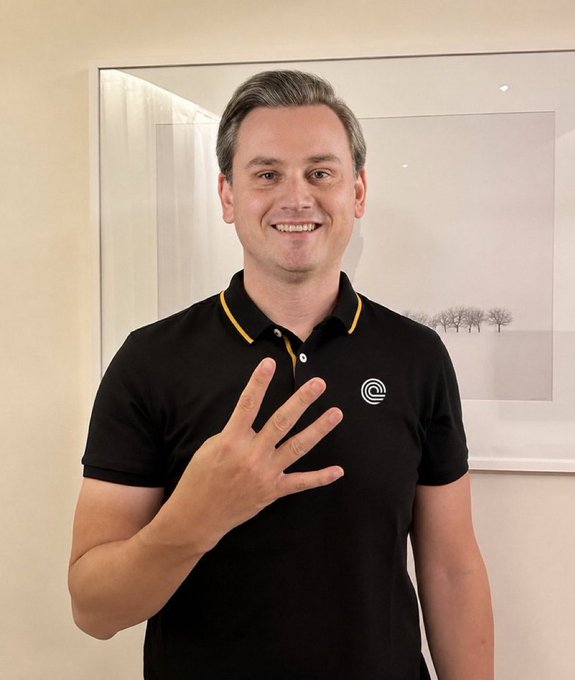 A smiling man wearing a black polo shirt with yellow trim and a circular logo, holding up his right hand showing five fingers, standing in front of a framed picture on a beige wall.