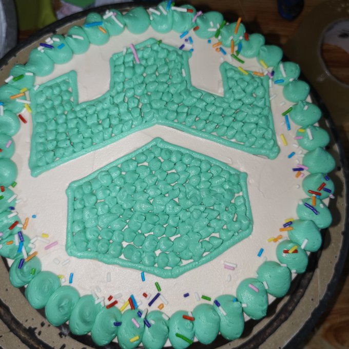 A homemade cake decorated with teal frosting in the shape of a crown emblem, bordered with piped frosting dots and rainbow sprinkles. The post celebrates Gamdom's giveaway, with the baker sharing their TRC20 wallet address for the crypto tip.
