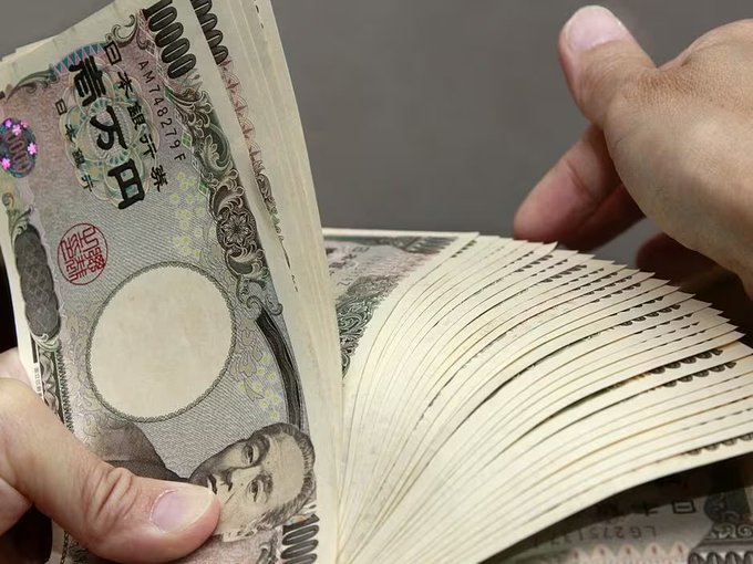 A pair of hands fans through a thick stack of Japanese 10,000 yen banknotes, prominently featuring the distinctive portrait and "10000" denomination. The visual of abundant yen currency reinforces the article's theme of cheap yen borrowing fueling carry trades, where investors leverage low-interest Japanese currency to invest in higher-yielding assets like cryptocurrencies.