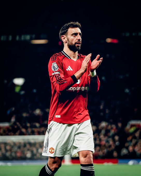 A Manchester United player wearing a red home kit with 'Snapdragon' sponsorship and the Premier League badge applauds the crowd with a serious expression during a night match.