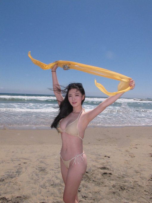 A young woman stands on a sandy beach in a beige bikini, holding a flowing yellow scarf above her head while waves roll in behind her under a clear blue sky. The image appears to be a casual, sunlit beach portrait with no visible text or numbers, simply capturing a relaxed seaside moment.